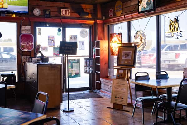 View of Darrell's restaurant entrance from inside, featuring a host stand, a 'Please Wait to Be Seated' sign, and window seating surrounded by neon beer signs and natural light from large front windows