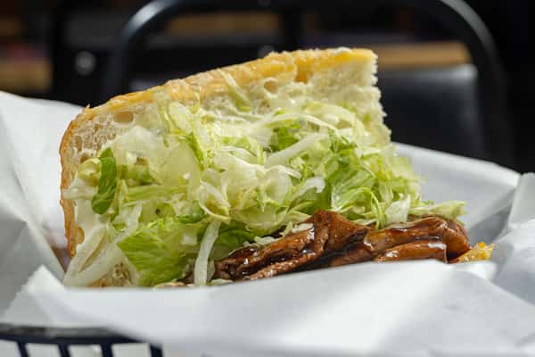 Close-up cross-section of a po'boy sandwich loaded with tender grilled meat in a tangy BBQ glaze, topped with shredded lettuce and served on a soft toasted bun.