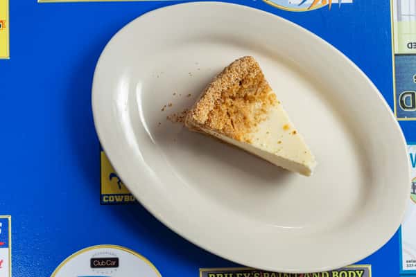 Top-down view of a plain slice of cheesecake with a graham cracker crust, served on a white plate atop a bright blue table at Darrell's.