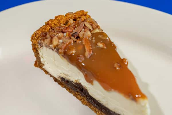 Decadent slice of Southern-style caramel pecan cheesecake with a graham cracker crust, rich chocolate fudge base, creamy cheesecake filling, and glossy caramel topping sprinkled with chopped pecans, served on a white plate.