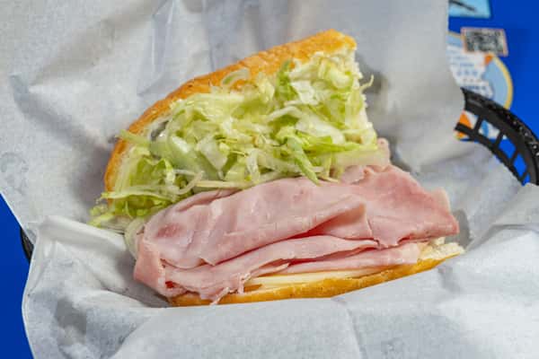 Open-faced ham po'boy sandwich with layers of sliced ham and shredded lettuce on a soft hoagie roll, served in a parchment-lined black basket.