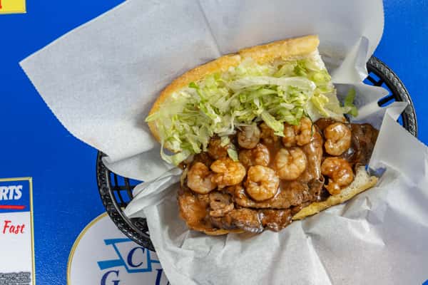 Overhead view of Darrell's surf and turf po'boy, featuring a generous layer of grilled steak and seasoned shrimp drenched in savory brown gravy, topped with shredded lettuce and served open-faced in a black basket lined with parchment paper on a blue tabletop
