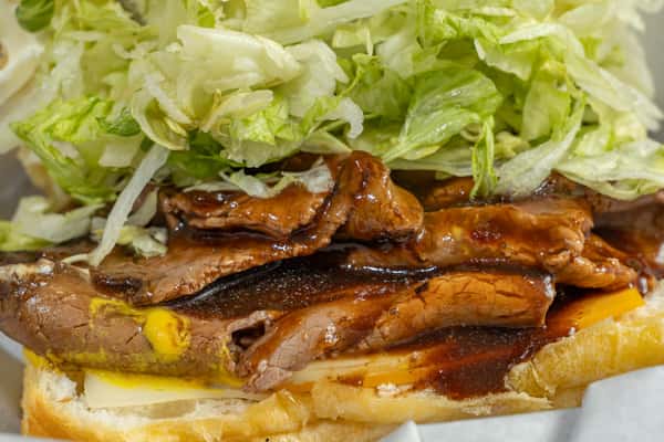 Extreme close-up of a steak po'boy showcasing grilled steak slices glazed with sauce, melted cheese, and shredded lettuce in a fresh sandwich roll.