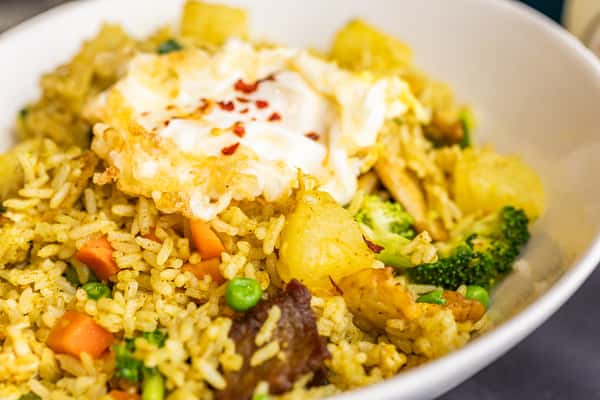 Fried rice with mixed vegetables, chunks of meat, and a crispy fried egg topped with chili flakes in a white bowl.