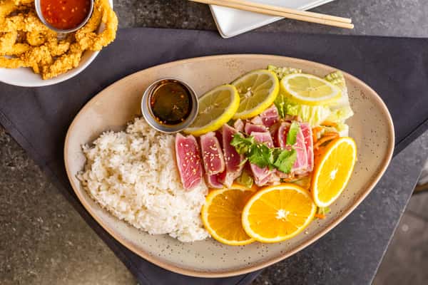 Seared ahi tuna with white rice, citrus slices, fresh greens, and soy dipping sauce on a beige plate.