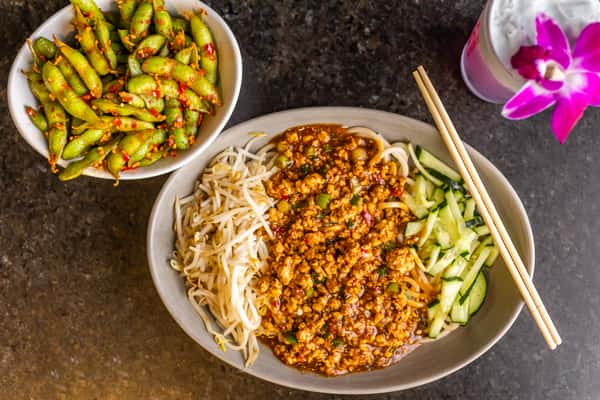 Bowl of noodles with spicy minced meat, bean sprouts, and cucumber, served with chili edamame and a drink garnished with an orchid.