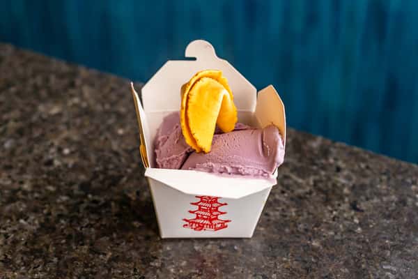 Scoops of purple ube ice cream topped with a fortune cookie, served in a classic Chinese takeout box.