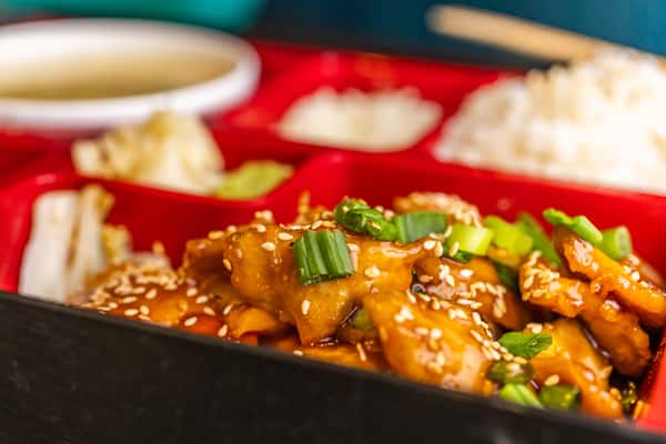 Sesame chicken topped with green onions in a bento box, served with rice, dumplings, wasabi, and pickled ginger.