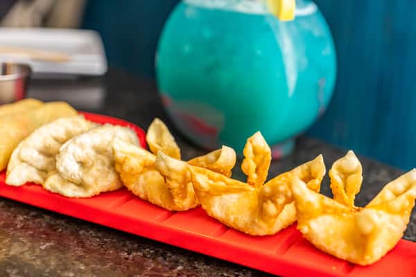 Golden fried wontons, crispy dumplings, and spring rolls served on a red tray beside a vibrant blue tropical cocktail.