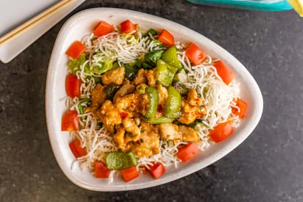 Crispy noodles topped with stir-fried chicken, bell peppers, lettuce, and diced tomatoes in a white triangular bowl.