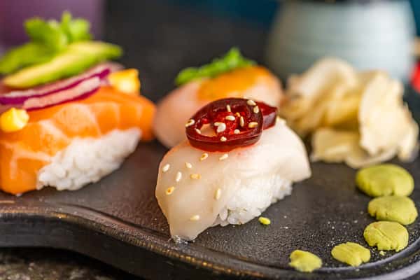 Assorted nigiri sushi topped with jalapeño, onion, and avocado, served with wasabi and pickled ginger on a black stone platter.
