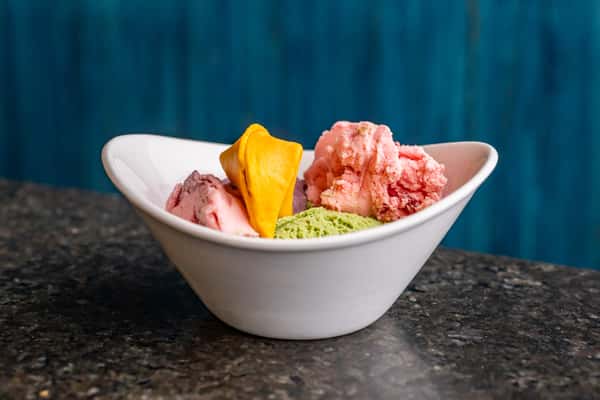 Trio of colorful ice creams with a dried mango slice, served in a white bowl against a blue background.