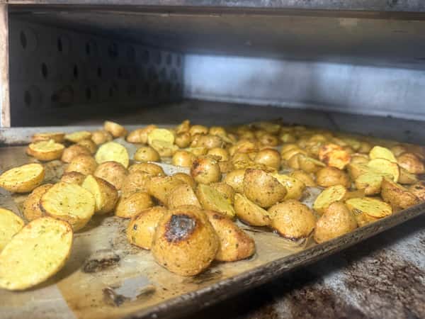 Golden roasted baby potatoes seasoned with herbs, baking on a metal tray inside a commercial oven—perfect as a rustic side dish or catering option.