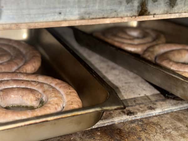 Coiled Italian sausages roasting in metal trays inside a commercial oven, slowly browning in their natural juices—perfect for hearty, rustic dishes or traditional catering menus.