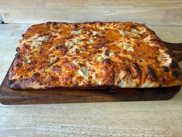 Rectangular pan-style pizza with a thick, crispy crust, generously topped with melted cheese and a creamy orange-hued sauce, served on a wooden paddle.