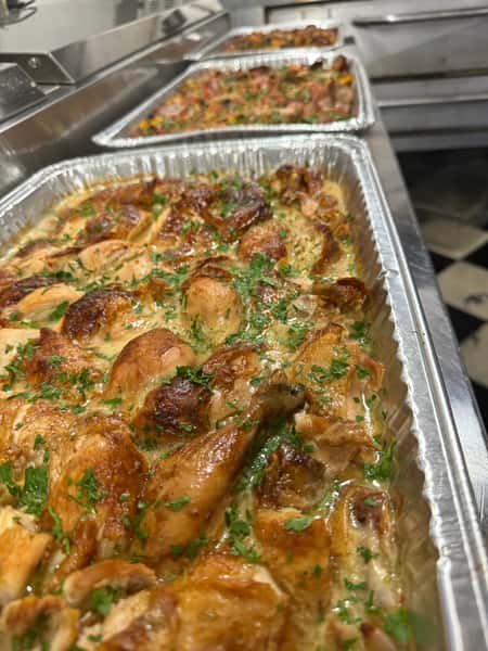 Front-facing view of multiple catering trays filled with roasted chicken in creamy herb sauce, garnished with fresh parsley, lined up in a commercial kitchen—ideal for large events or buffet-style service.