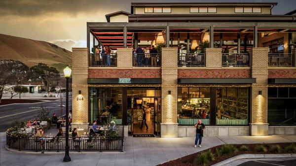 Modern restaurant exterior with outdoor patio seating, rooftop dining, and guests enjoying an upscale evening atmosphere.