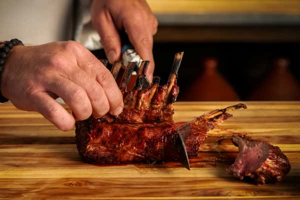 Chef carving a slow-roasted rack of meat on a wooden board in an open restaurant kitchen.
