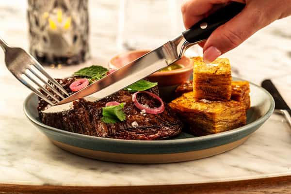 Premium grilled steak with chimichurri, pickled onions, and crispy layered potatoes being sliced tableside.