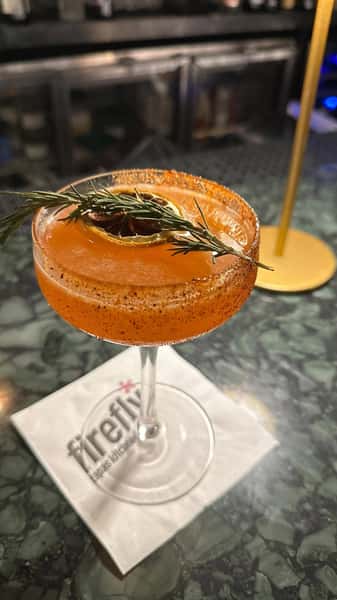 A vibrant cocktail in a coupe glass, with an orange slice floating on top, garnished with a fresh rosemary sprig. The glass has a spicy‑rim sugar or salt blend, the drink is a rich amber/orange color, and it rests atop a white napkin on a marble or patterned bar counter, with dim bar shelves blurred in the background.