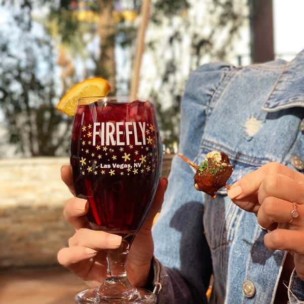 Close-up of a red sangria with an orange garnish in a branded Firefly Las Vegas glass, paired with a bite-sized tapas skewer, held by a person in a denim jacket at a sunlit outdoor patio.
