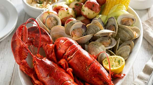The image features a vibrant seafood boil with a whole lobster as the centerpiece, surrounded by clams, red potatoes, corn on the cob, and sausage. Everything is coated in a seasoned, buttery sauce, giving the dish a rich, glossy look. The colors are bold and appetizing, capturing a hearty, coastal feast perfect for sharing.