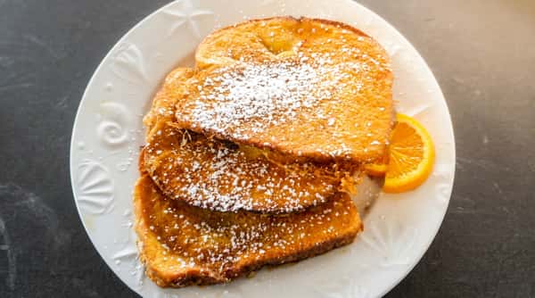 A stack of golden-brown pancakes dusted with powdered sugar sits on a plate, served with a side of syrup. The pancakes look fluffy and warm, perfect for a comforting breakfast.