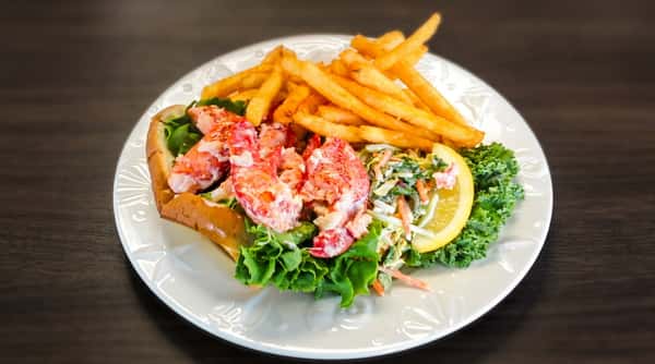 A fresh lobster roll is served on a bed of greens with a side of crispy golden fries and a lemon wedge. The lobster meat looks tender and generously portioned, creating a classic coastal favorite.