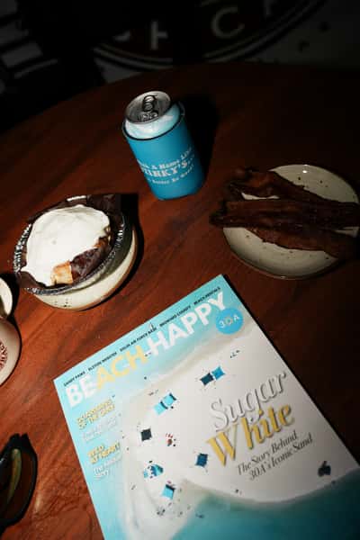 Table scene with dessert topped with whipped cream, crispy bacon, and a canned drink in a Stinky's koozie, next to a Beach Happy 30A magazine on a wooden table.