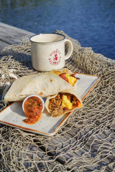Breakfast burrito with eggs, bacon, and sausage on a plate with salsa, beside a Stinky's mug on a dock by the water.