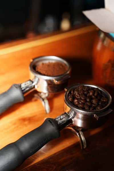 Two espresso portafilters—one filled with freshly ground coffee and the other with whole coffee beans—resting on a wooden counter, ready for brewing.