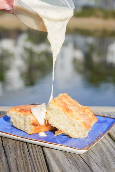 Creamy gravy being poured over a warm biscuit on a blue plate, set on a wooden dock beside calm water.