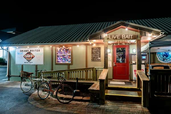 Stinky's Bait Shack entrance at night with a "Live Bait" sign, string lights, red door, and bicycles parked outside.