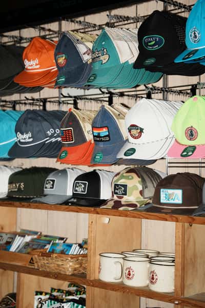 Display wall of colorful branded hats and merchandise from Stinky's Fish Camp and related coastal brands, arranged on racks above souvenir mugs and retail items in a gift shop area.
