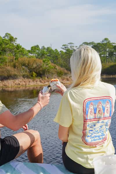 Two people sitting by the water sharing a wrapped breakfast burrito, wearing a Stinky's shirt with coastal trees in the background.