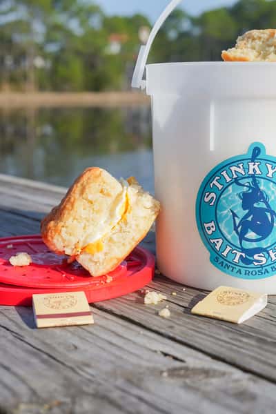Cheddar biscuit and butter beside a Stinky's Bait Shack bucket on a dock overlooking calm coastal water.