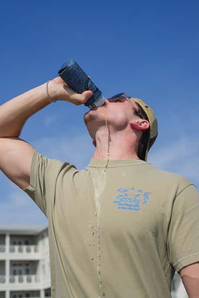 Person drinking from a can in a Stinky's Fish Camp koozie, wearing a Stinky's shirt outdoors under a clear blue sky.
