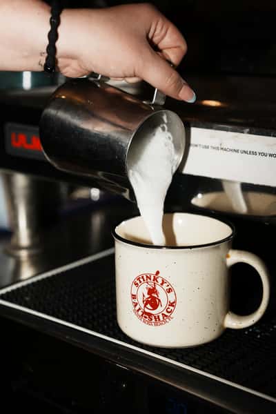 Milk being poured into a Stinky's Bait Shack branded mug at an espresso machine, preparing a hot coffee drink.