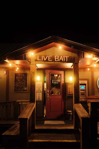Entrance to a bait and tackle shop at night with a glowing "Live Bait" sign, string lights, and an open red door.