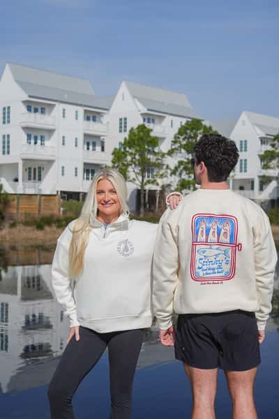 Two people modeling Stinky's branded sweatshirts outdoors by a waterfront, with white coastal buildings in the background. One person faces forward wearing a cream quarter-zip with a small chest logo, while the other faces away showing a colorful Stinky's graphic on the back.