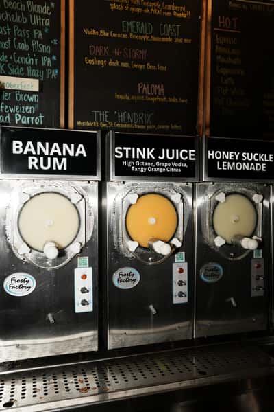 Frozen drink machines at a bar featuring flavors Banana Rum, Stink Juice, and Honey Suckle Lemonade, with a chalkboard cocktail menu behind them.