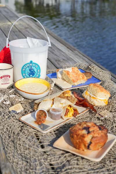 Coastal dockside breakfast spread from Stinky's Bait Shack featuring breakfast tacos, biscuits, grits, and a blueberry muffin with water in the background.