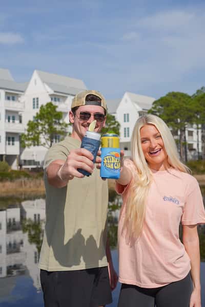 Two people toasting drinks in Stinky's Fish Camp koozies by a waterfront with white coastal buildings in the background.