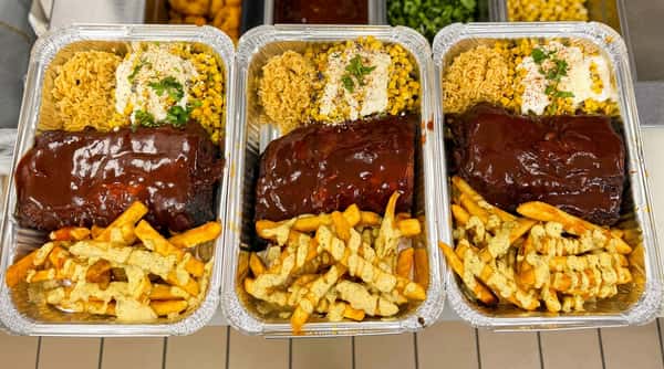Three foil containers with BBQ ribs, french fries with a creamy sauce, seasoned rice, and corn with cotija cheese