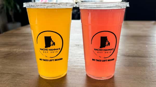 Two plastic cups with lids on a wooden table, one filled with yellow liquid and the other with orange-red liquid. Both cups have a black logo with "NACHO MAMMA'S EST 2010 NO TACO LEFT BEHIND" and an outline of Rhode Island.