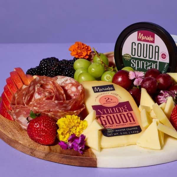 Charcuterie board with Marieke Young Gouda, Gouda Spread, salami, fruit, and edible flowers on a lavender background.