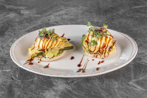 Two servings of avocado toast topped with sliced avocado, fresh herbs, and a drizzle of dark sauce, beautifully presented on a white plate.
