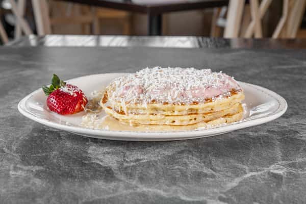 Stack of golden pancakes dusted with powdered sugar, served on a white plate with fresh strawberries on the side.