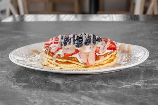 Stack of golden pancakes topped with fresh strawberries, blueberries, whipped cream, and a dusting of powdered sugar, served on a white plate.
