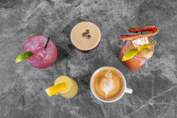 Overhead view of assorted colorful beverages, including a latte with heart-shaped latte art, iced coffee, and refreshing fruit drinks on a gray tabletop.